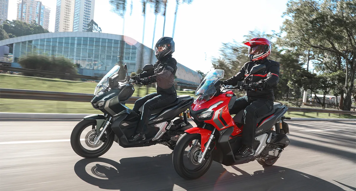 Mulher em Honda ADV preta com capacete preto e homem em Honda ADV vermelha de capacete vermelho em rua de cidade