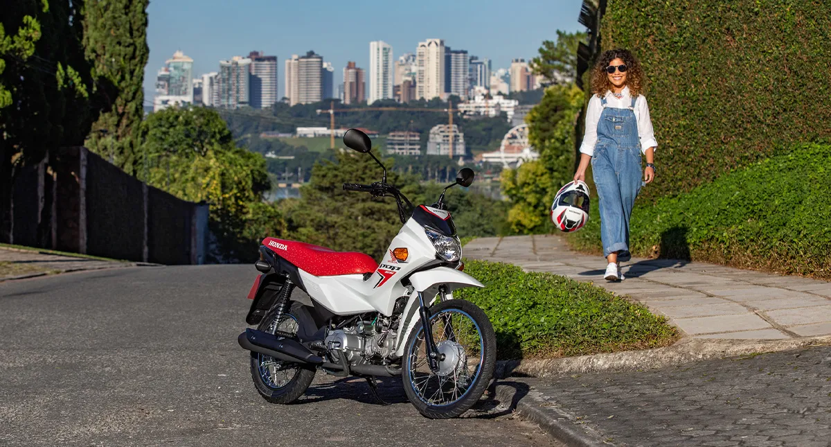 Mulher sorridente com capacete em mãos indo ao encontro de sua motocicleta Honda Pop