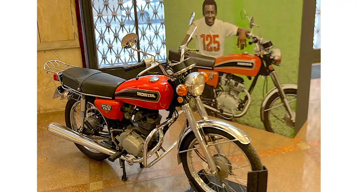 Motocicleta Honda 125 em exibição com um painel informativo ao fundo